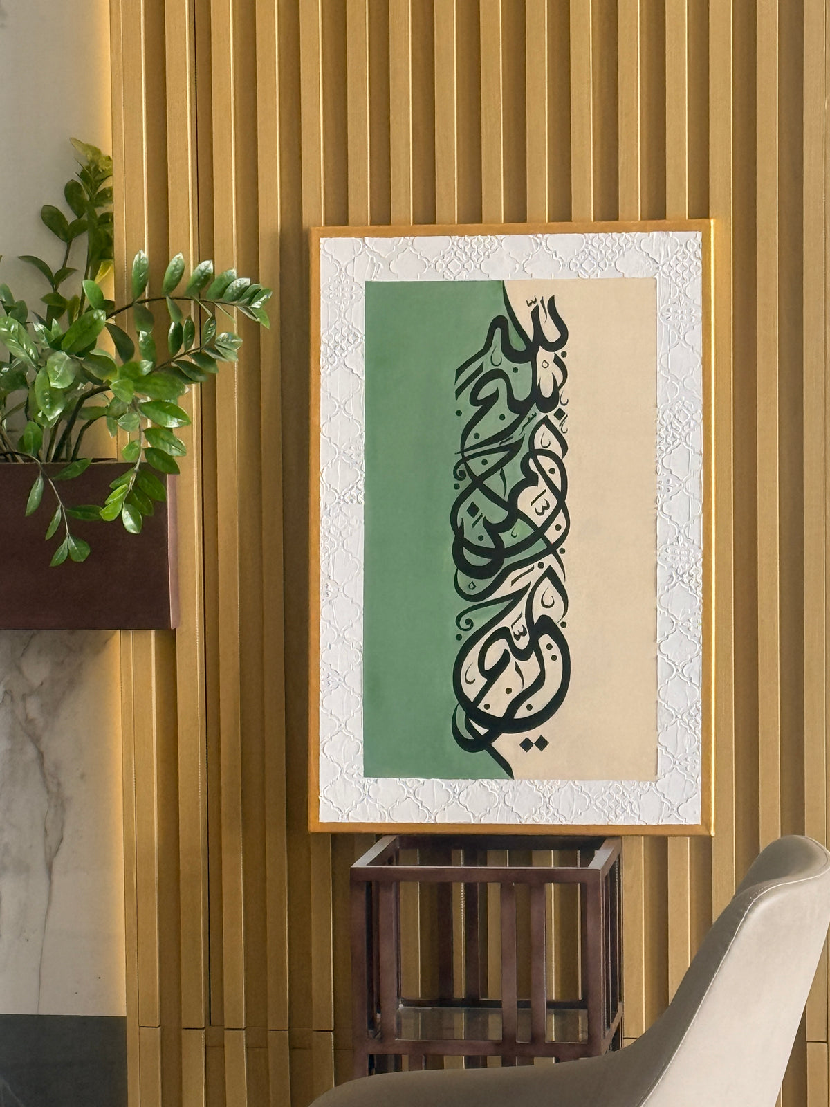 Framed Islamic calligraphy wall art hanging on a slatted wood wall, featuring black Arabic script over a split green and beige background with a white textured border. 