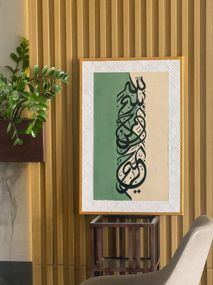 Framed Islamic calligraphy wall art hanging on a slatted wood wall, featuring black Arabic script over a split green and beige background with a white textured border. 